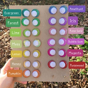 Könnte beinhalten: Eine Sammlung glitzernder Bastelaugen in verschiedenen Farbtönen, darunter Grün, Lila, Rosa und Orange. Jedes Auge hat eine weiße Mitte und ist mit einem Farbnamen wie "Evergreen" und "Amethyst" beschriftet. Die Augen sind auf einem Karton angeordnet.
