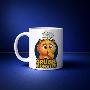 Può includere: Tazza in ceramica bianca con l'illustrazione di un mostro dei cartoni animati. Il mostro arancione ha le corna e un'espressione preoccupata. La tazza presenta il testo "GRÜBEL MONSTER" in giallo e bianco. Una nuvoletta con punti interrogativi e spirali fluttua sopra la testa del mostro.