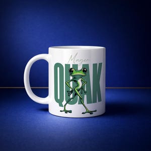 May include: White ceramic mug featuring a green cartoon frog with the word "QUAK" in large green letters. The word "Magen" is written above the word "QUAK" in a script font. The frog is standing with its arms crossed.