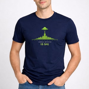 Peut inclure: T-shirt bleu marine avec un motif vert d'OVNI au-dessus d'un indicateur de signal. Le texte "ANOMALIE DÉTECTÉE" et "1,6 GHz" sont en dessous. Le t-shirt est de style col rond.