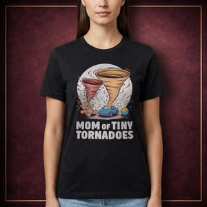 Può includere: T-shirt nera con una grafica raffigurante due tornado dei cartoni animati, un orsacchiotto, un'auto blu e una palla. Il testo "MOM of TINY TORNADOES" è stampato in bianco sotto.