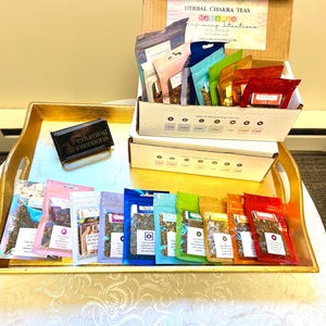 Op de afbeelding: Een gouden dienblad met zeven rijen kleine, kleurrijke zakjes met kruidenthee-mengsels. De zakjes zijn gelabeld met de namen van de zeven chakra's. Het dienblad staat op een witte tafel. De tekst op de doos luidt "Herbal Chakra Teas Perfuming Intentions".