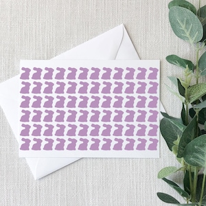 May include: A white greeting card with a pattern of lavender bunny silhouettes. The card is placed on top of a white envelope. Green leafy sprigs are in the background. The card is suitable for Easter or spring celebrations.