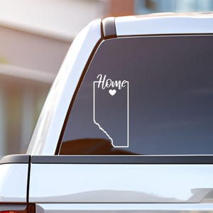 May include: White vinyl decal of the outline of the province of Alberta with the word "Home" and a small heart above it. The decal is on the back window of a silver pickup truck.