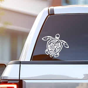 May include: A white vinyl decal of a sea turtle applied to the rear window of a vehicle. The turtle shell features a detailed cutout pattern of hibiscus flowers and tropical leaves, creating a decorative graphic design on the dark glass surface.