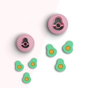 May include: Pink avocado-shaped cookie cutters and avocado-shaped food decorations. The cutters are pink with a center cutout. The avocado decorations are green with a yellow center.