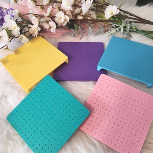 May include: Five colorful, square, perforated mats in yellow, purple, blue, teal, and pink. Each mat has a slightly raised edge on one side. The mats are arranged on a white, fluffy surface, with floral accents in the background.