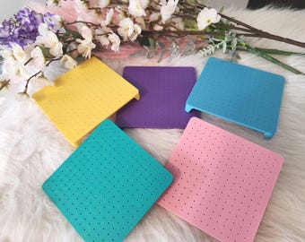 May include: Five colorful, square, perforated mats in yellow, purple, blue, teal, and pink. Each mat has a slightly raised edge on one side. The mats are arranged on a white, fluffy surface, with floral accents in the background.