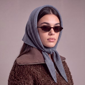 May include: A blue knitted headscarf tied under the chin, worn with oval-shaped tortoiseshell sunglasses and a brown textured zip-up jacket with a ribbed collar. The headscarf features a fine waffle-like knit pattern and pointed ends.