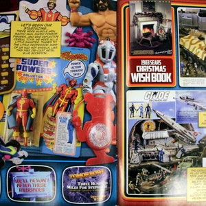 May include: A vintage magazine spread featuring action figures, a calculator, and a Christmas Wish Book. The toys include a Wonder Woman doll, Super Powers Collection figures, and G.I. Joe sets. The text includes "Super Powers Collection" and "1983 Sears Christmas Wish Book."