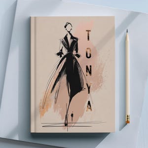 May include: A beige notebook with a fashion illustration on the cover. The illustration is a black sketch of a woman in a coat, with the name "TONYA" in gold lettering. A pencil rests beside the notebook.