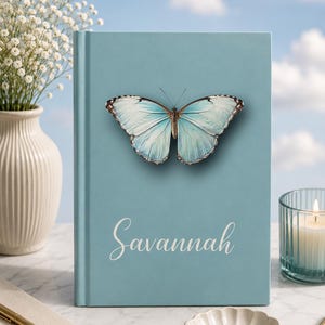 May include: A light blue journal with a butterfly illustration and the name "Savannah" in script. The butterfly has blue and white wings. A lit candle, a vase of white flowers, and a pen are also visible.