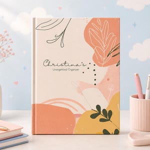 May include: A hardcover journal with a floral design in peach, yellow, and green. The cover reads "Christina's Unorganized Organizer" in script. The journal is on a desk with pens, a vase of flowers, and other notebooks.
