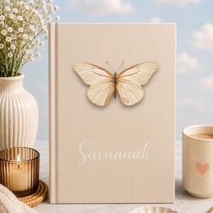 May include: A beige journal with a butterfly illustration and the name "Savannah" in script. The journal is surrounded by a candle, a mug with a heart, and a small dish with paper clips. A vase of white flowers is in the background.