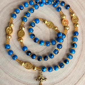 May include: A rosary and matching bracelet set. The rosary features speckled blue beads, gold-colored spacers, and a gold-tone crucifix. The bracelet has similar blue beads and gold accents. The set is displayed on a light wood surface.