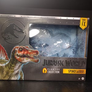 Może przedstawiać: Figurka Spinosaurusa z kolekcji Jurassic World Hammond w oryginalnym opakowaniu. Pudełko zawiera ilustrację Spinosaurusa w odcieniach czerwieni, brązu i zieleni. Opakowanie zawiera tekst "Jurassic World" i "Spinosaurus".