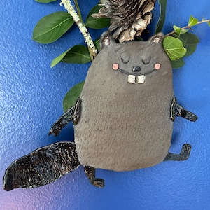 May include: A handcrafted ceramic wall hanging shaped like a stylized beaver with a dark gray textured body, a glossy black tail, two white front teeth, and small pink circular cheeks, positioned against a blue background with green leaves and a pinecone.