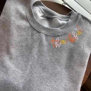 Peut inclure: Sweat-shirt gris avec le mot "thankful" écrit en cursive orange, rose et jaune.