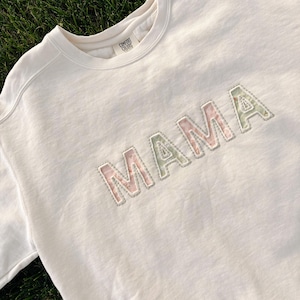 Könnte beinhalten: Cremefarbenes Sweatshirt mit dem Wort "MAMA" auf der Brust gestickt. Die Buchstaben sind mit einem floralen Muster in Rosa- und Grüntönen gefüllt, umrandet von einer grauen Naht. Das Sweatshirt ist aus weichem Material.