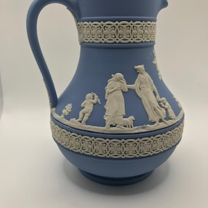 May include: A blue ceramic pitcher with a white handle and decorative trim. The pitcher features white relief figures and floral patterns around the top and base. A classic piece for serving beverages.