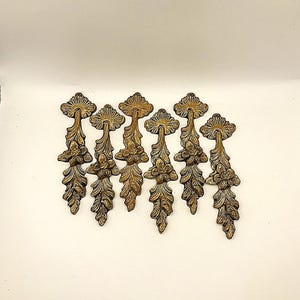 Peut inclure: Sept poignées décoratives antiques en laiton avec des motifs floraux et de feuilles. Chaque poignée présente un dessus en forme d'éventail, une fleur centrale et un motif de feuilles en cascade. Les poignées sont disposées sur une surface blanche.