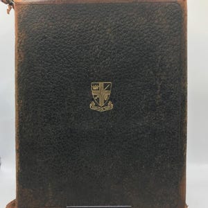 May include: A vintage, dark brown leather-bound book with a gold emblem on the front. The book shows signs of wear, with a distressed appearance and a slightly torn top edge. The emblem features a crest design.
