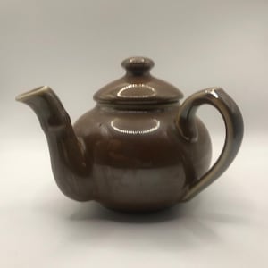 Könnte beinhalten: Eine braune Keramik-Teekanne mit Deckel und Henkel. Die Teekanne hat einen abgerundeten Körper und einen Ausguss. Der Griff und der Ausguss haben eine hellere braune Farbe. Die Teekanne steht vor weißem Hintergrund.