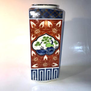 May include: A rectangular ceramic vase with a detailed design. The vase features a red background with white and gold floral patterns. A central panel depicts a landscape scene with green foliage and blue accents. The top has a blue and gold checkered pattern.