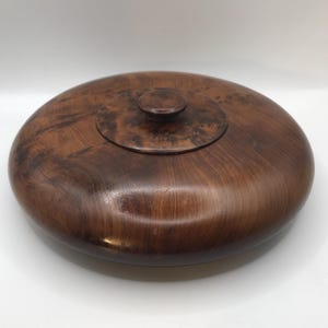 May include: A handcrafted wooden bowl with a lid. The bowl is oval-shaped and made of dark brown wood with a natural grain pattern. The lid has a small, round knob for easy opening. The bowl is approximately 10 inches in diameter.