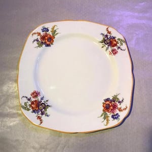 May include: Square white plate with a scalloped edge and floral design. The plate features clusters of red, orange, purple, and blue flowers in each corner, with a gold-coloured trim.