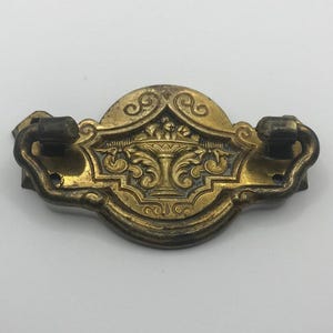 Op de afbeelding: Osiersierlijke messing ladeknop met een centraal bloemmotief en gebogen handgrepen. De antieke hardware heeft een gedetailleerd, symmetrisch patroon en een warme, verouderde afwerking, ideaal voor restauratie van antieke meubels.