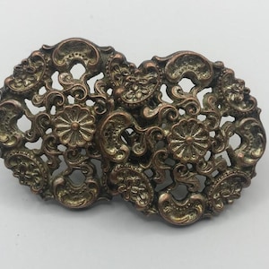 May include: An ornate, vintage-style metal buckle with a bronze finish. The buckle features an openwork design with floral and scroll details. The two halves are connected, creating a symmetrical shape. A decorative accessory.