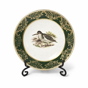 May include: A round ceramic plate featuring a central illustration of a sandpiper bird standing on rocks. The plate has a wide dark green border decorated with a gold lattice pattern and floral motifs, finished with a thin gold rim around the edge.