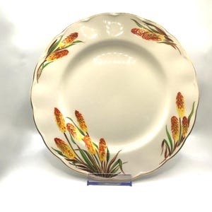 May include: Cream-coloured ceramic plate with a scalloped edge and a gold rim. The plate is decorated with stylized yellow and orange floral designs with green leaves. A decorative piece for the kitchen or dining room.