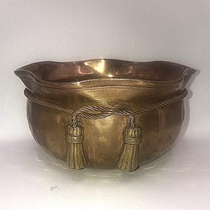 Może przedstawiać: Miska w kolorze mosiądzu z falistym brzegiem i wzorem sznurka pośrodku. Dwa chwosty zwisają ze sznurka. Miska ma zaokrąglony kształt i błyszczące wykończenie.