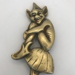 May include: A brass figurine depicting a mischievous creature with pointed ears and a sharp chin, sitting atop a rounded, textured mushroom-shaped base. The metal has a polished gold-toned finish with visible dark patina in the recessed areas.
