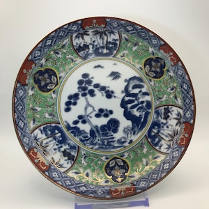 May include: A round ceramic plate featuring a central blue and white landscape illustration of pine trees and mountains. The border is decorated with green, blue, and red floral patterns, gold accents, and geometric shapes in a circular arrangement.