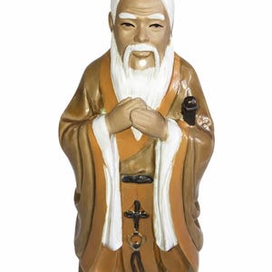 May include: A ceramic figurine of an elderly person with a long white beard and hair, wearing a brown and tan robe with white trim. The figure has hands clasped at the chest and features a small black accessory hanging from the waist area.