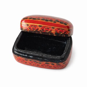 Könnte beinhalten: Eine rechteckige Lackschatulle mit Klappdeckel, die eine glänzend rote Außenseite mit einem schwarzen Blumenmuster aufweist. Das Innere der Schatulle ist einfarbig schwarz lackiert und der Deckel ist leicht angehoben, was das leere Fach freigibt.