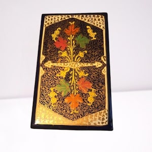 Pode incluir: Uma caixa decorativa retangular preta com um design floral. A caixa apresenta uma borda dourada e um padrão detalhado. O design central inclui folhas coloridas em vermelho, laranja e verde, com detalhes dourados. A caixa tem um acabamento brilhante.