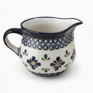 Op de afbeelding: Een keramieken roomkannetje met een bolvormig lichaam en een gebogen handvat. Het crèmekleurige oppervlak is versierd met een herhalend patroon van donkerblauwe bloemen met oranje harten, afgewisseld met kleine groene bladmotieven en een blauwe rand.
