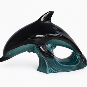 Könnte beinhalten: Eine glänzende Keramikfigur eines Delfins in Tauchpose, mit einem glatten schwarzen Oberkörper und einem tiefen blaugrünen Unterteil. Der Delfin ist über einer stilisierten, geformten Wellenbasis in passender blaugrüner Glasur positioniert.
