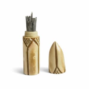 Peut inclure: Un étui à aiguilles cylindrique sculpté dans un matériau de couleur crème avec des accents bruns, présentant un motif quadrillé sur le dessus. L'étui est ouvert, révélant un groupe d'aiguilles en métal argenté, avec un capuchon pointu assorti.