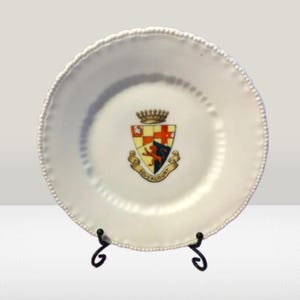 May include: White ceramic plate with a raised edge and a coat of arms design in the center. The coat of arms features a lion, a crown, and the text "Dovercourt". The plate is on a black metal stand.