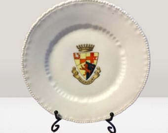 May include: White ceramic plate with a raised edge and a coat of arms design in the center. The coat of arms features a lion, a crown, and the text "Dovercourt". The plate is on a black metal stand.