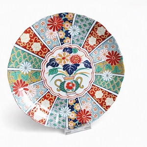 May include: A round ceramic plate featuring a central medallion with a vase of flowers surrounded by radiating panels. The design includes red, green, blue, and white floral patterns, geometric shapes, and scalloped edges on a white background.