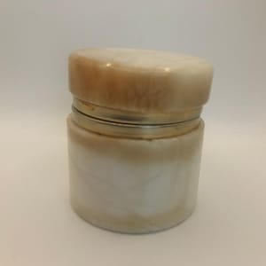 Puede incluir: Un pequeño tarro redondo con tapa. El tarro está hecho de un material blanco cremoso con vetas marrones y beige. La tapa es de un color y forma similares. Una banda plateada rodea el centro del tarro.