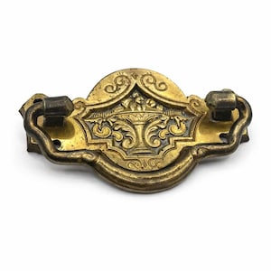 Peut inclure: Une poignée de tiroir en laiton antique présentant un motif central d'urne en relief entouré de volutes complexes. La quincaillerie comprend une poignée pivotante incurvée fixée à une plaque décorative avec deux trous de montage latéraux.