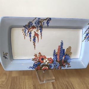 Puede incluir: Bandeja rectangular de cerámica con borde azul claro y centro blanco. La bandeja está decorada con motivos florales y de pájaros en rojo, azul y dorado. La bandeja tiene asas en cada extremo.