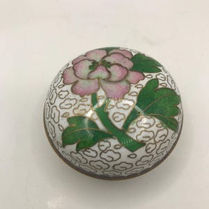 Pode incluir: Uma caixa de bugigangas redonda e decorativa com um design floral. A tampa apresenta uma flor rosa e verde com folhas verdes sobre um fundo branco com um padrão de nuvens. A caixa tem uma borda dourada.